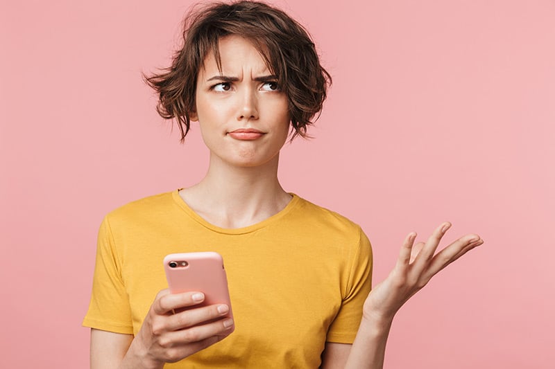 Confused woman posing isolated over pink wall background using mobile phone.